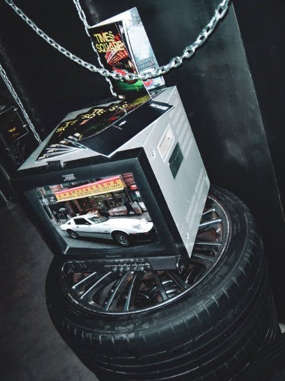 A member''s Chinatown Night Out frame on one of the four screens. Plinths built from wheels and stacked tires rather than gallery pedestals, because the work is about cars and the room should not pretend otherwise